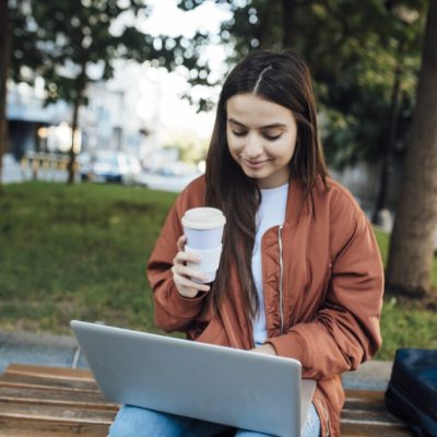 Girl with coffee cup and using laptop while sitting on bench.