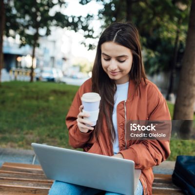 Girl with coffee cup and using laptop while sitting on bench.